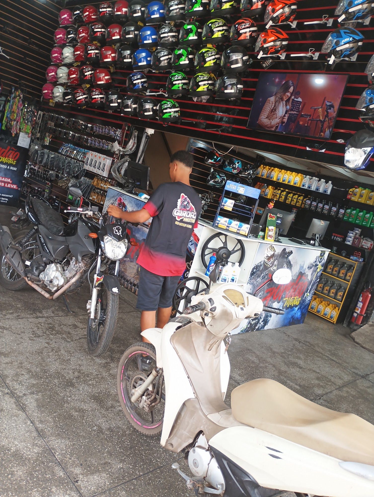 Atendente com camiseta Gordão no balcão de peças, com moto e scooter em primeiro plano