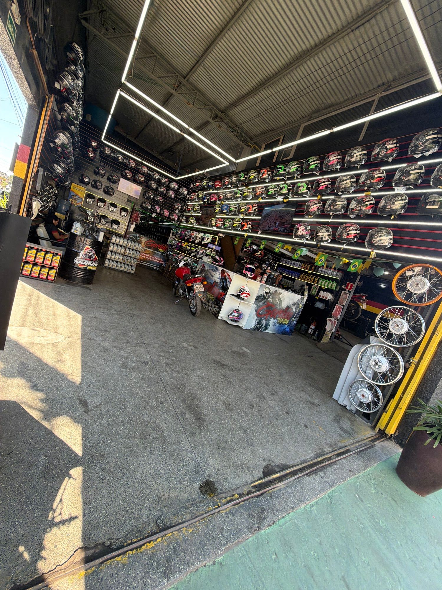 Fachada da Gordão Motos com a porta aberta: salão com paredes de capacetes iluminadas, balcão ao fundo e rodas de moto expostas